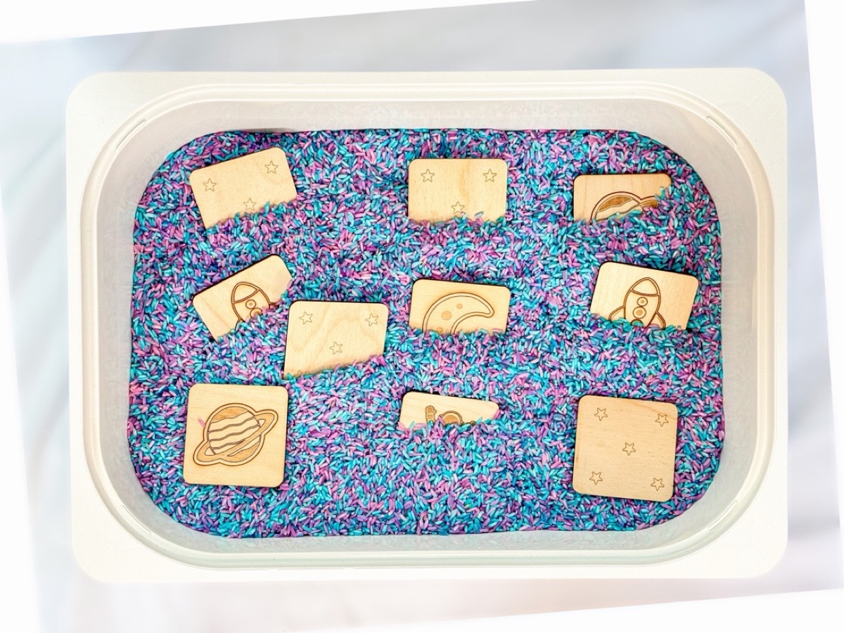 Wooden space tiles tucked into a sensory bin of purple and teal rice
