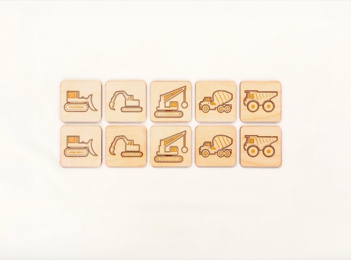10-piece neutral-colored construction vehicle memory match set on white, featuring five matching pairs of laser-engraved bulldozer, excavator, crane, cement mixer, and dump truck designs on neutral Baltic birch tiles. On a flat white background