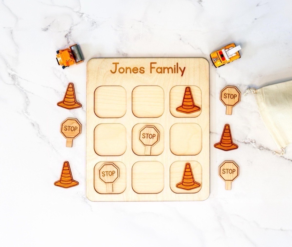 Personalized construction tic tac toe board engraved 'Jones Family' with traffic cone and stop sign playing pieces mid-game, toy work trucks and a muslin drawstring bag on the white marble surface