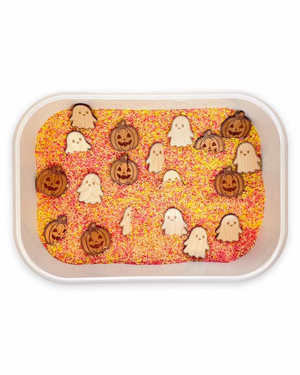 Nineteen wooden counters, eleven pale maple ghosts and eight reddish cherry jack-o'-lanterns, spread across orange, pink, and yellow dyed rice in a white rectangular tub, shown whole on a white background